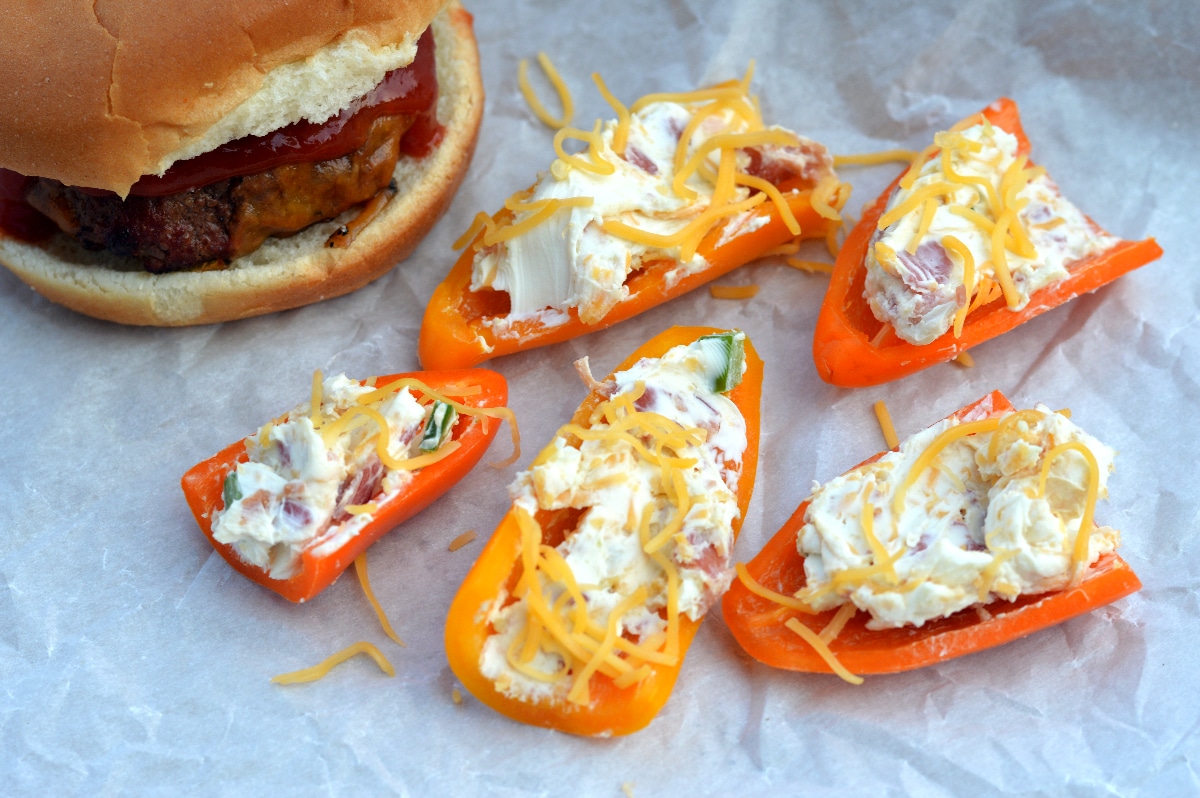 A photo of 5 stuffed mini pepper poppers next to a hamburger. Garnished with shredded cheddar cheese
