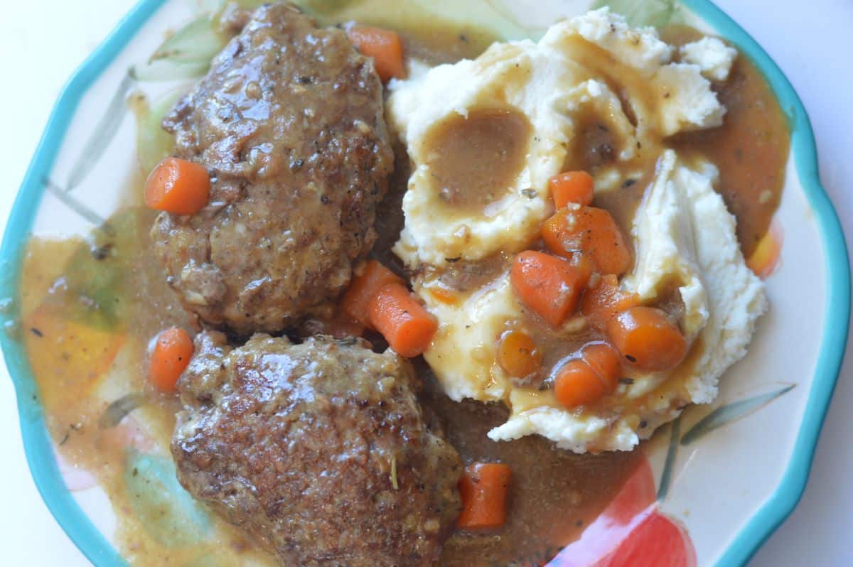 A photo of 2 hamburger steaks with diced carrots and gravy. Served on a plate with mashed potatoes.