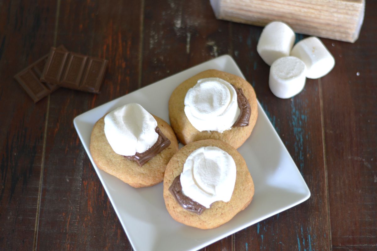 photo of 3 s'more cookies on a white plate, garnished with a chocolate square and melty marshmallow